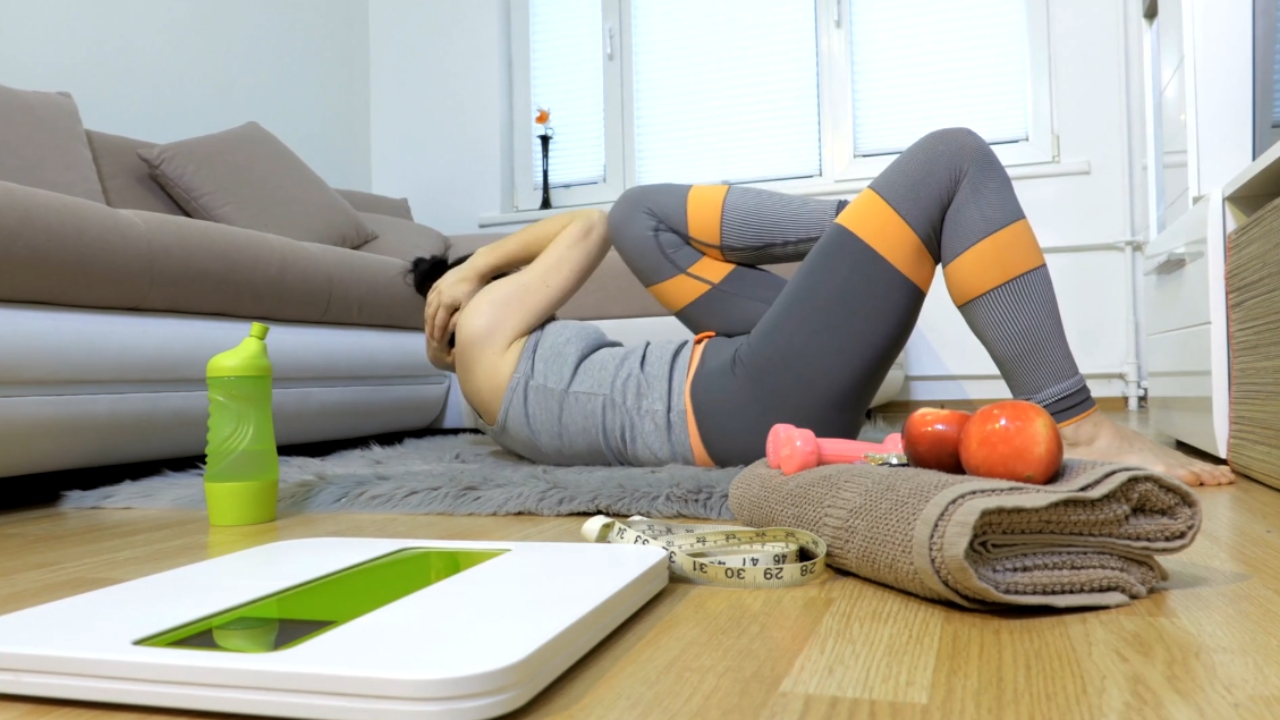 A woman exercises on the floor beside a scale, water bottle, and measuring tape.