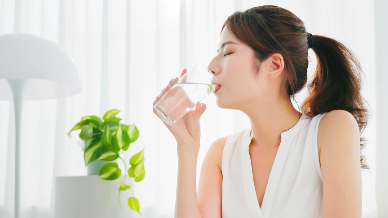 A woman drinks a glass of water beside a bright window.