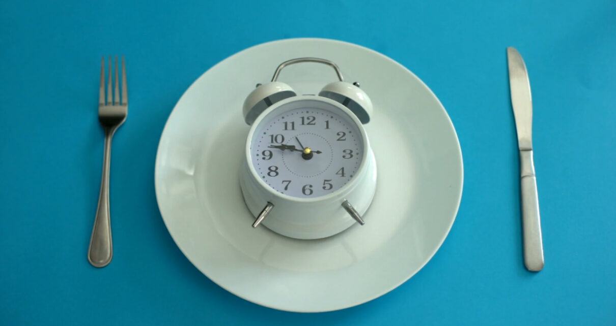 A white alarm clock sits on a plate between a fork and knife against a blue background.