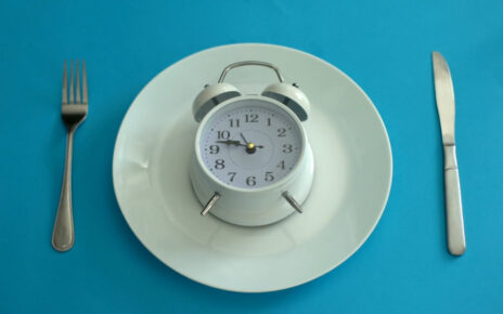 A white alarm clock sits on a plate between a fork and knife against a blue background.