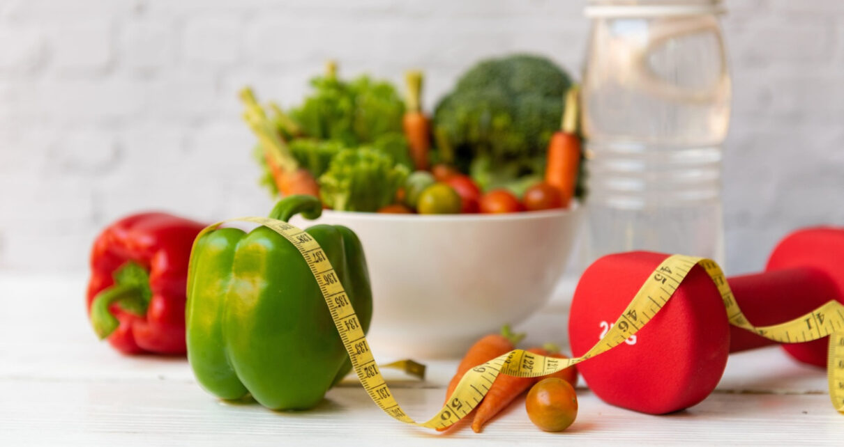 Fresh vegetables, a yellow measuring tape, and red dumbbells on a white table.