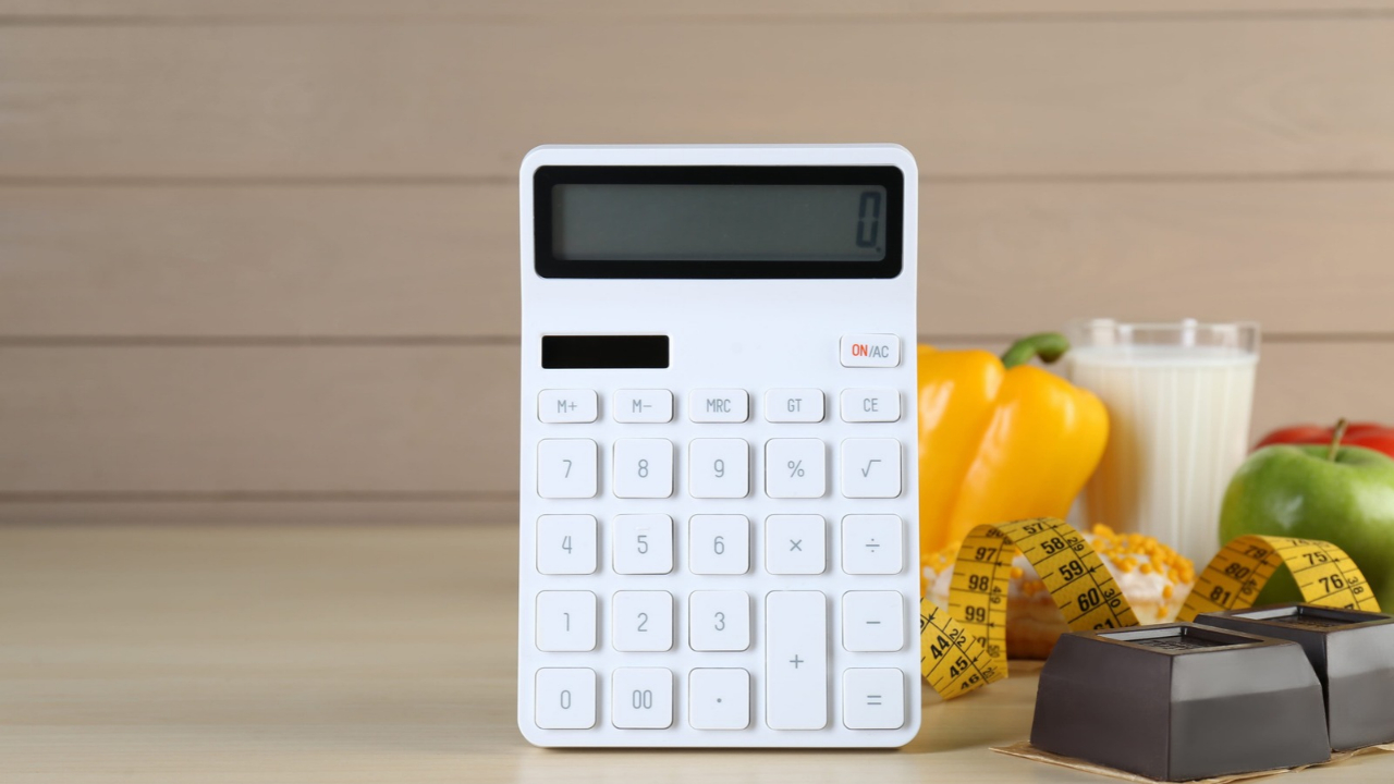 A calculator beside fresh vegetables, fruit, a glass of milk, and a measuring tape.
