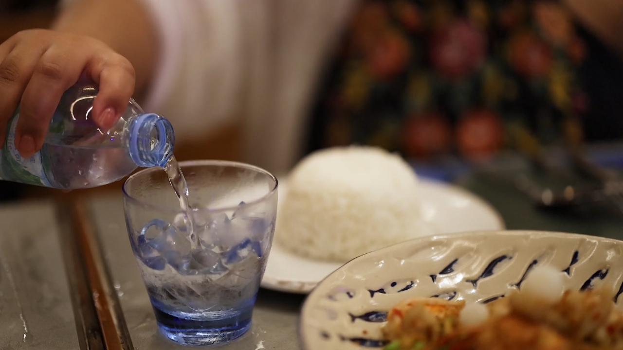 A person pours water into a glass beside a plate of food.