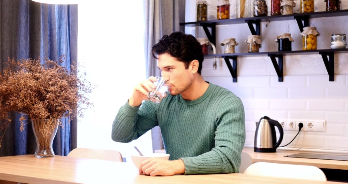 A man drinks water between bites at a kitchen table.