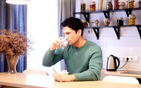 A man drinks water between bites at a kitchen table.