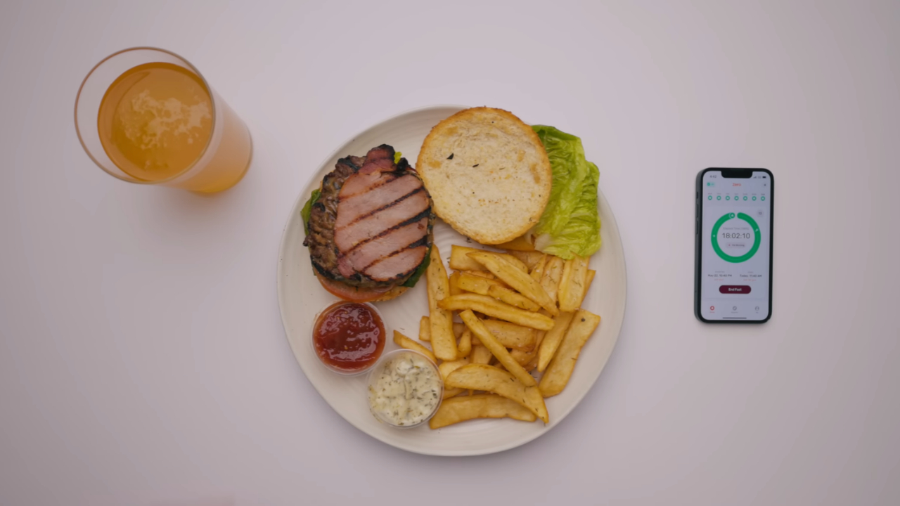 A burger with fries and dipping sauces sits beside a glass of juice and a phone with a fasting app.