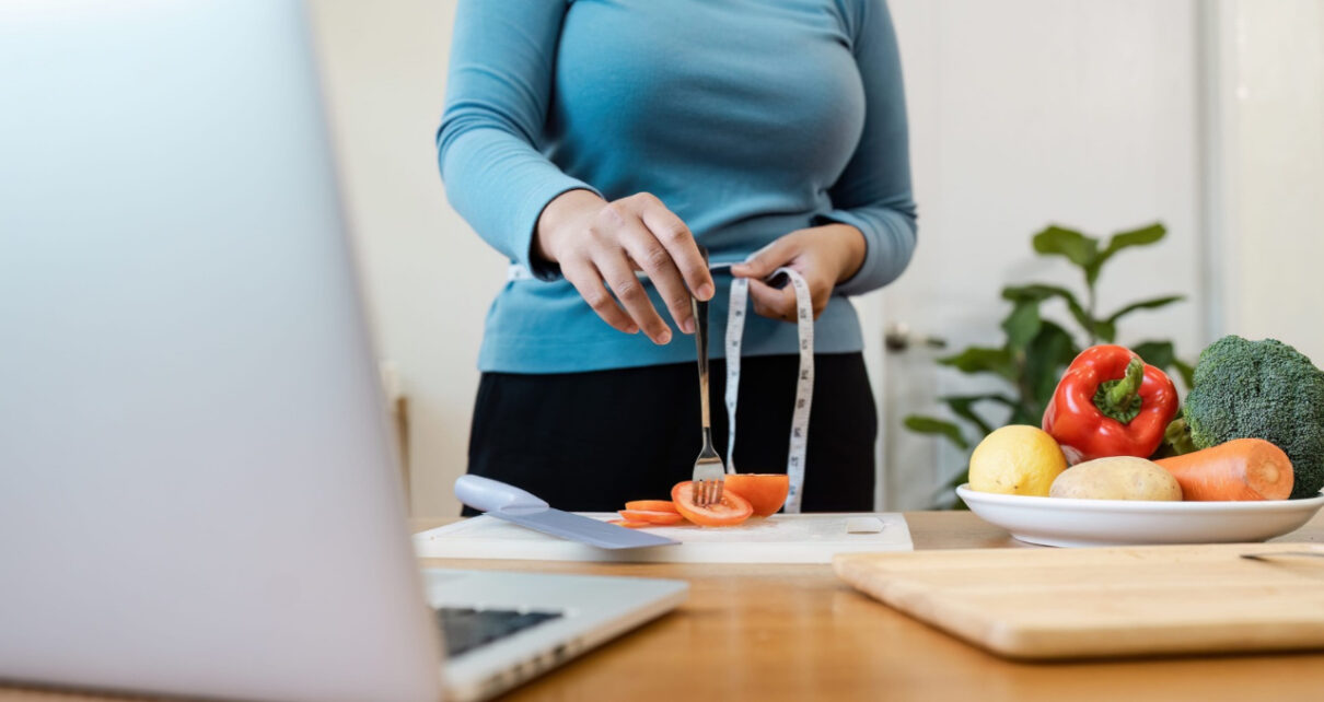 A woman measures her waist beside a laptop and fresh vegetables on a kitchen counter.