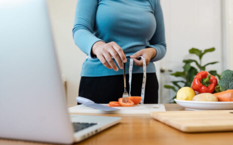 A woman measures her waist beside a laptop and fresh vegetables on a kitchen counter.