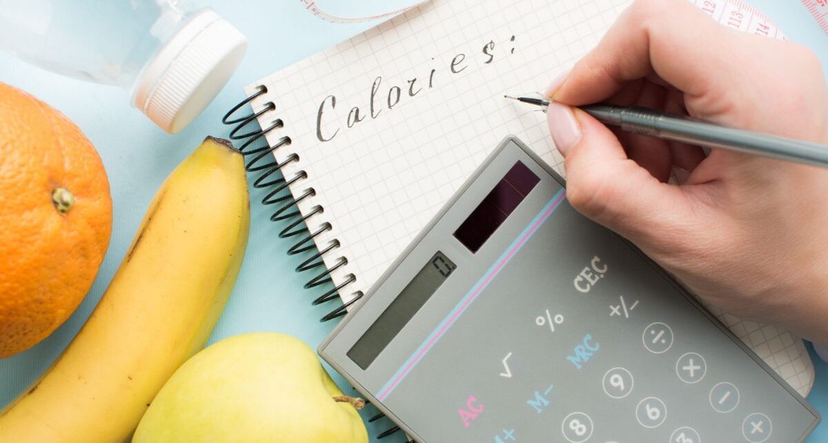 Woman calculating calories in paper notebook, calculator, bottle of water, measuring tape and fruits on blue background. Healthy eating concept - calculate daily nutrition intake.